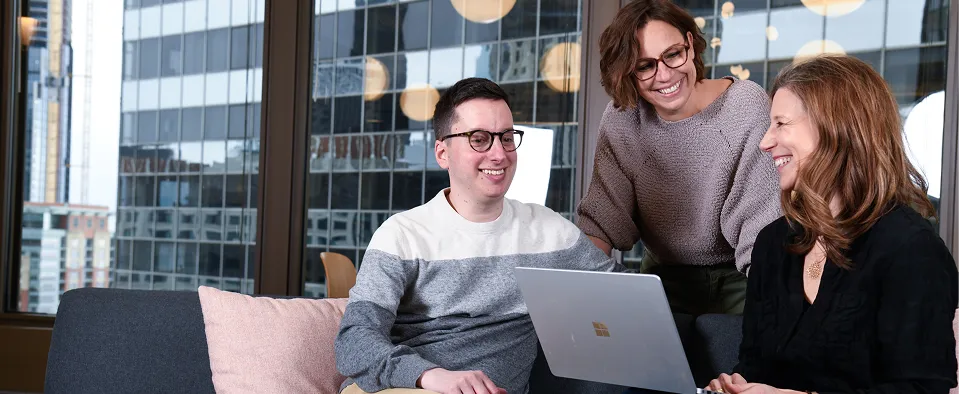 Three coworkers smiling and collaborating around a laptop in a modern office with city buildings visible through large windows.