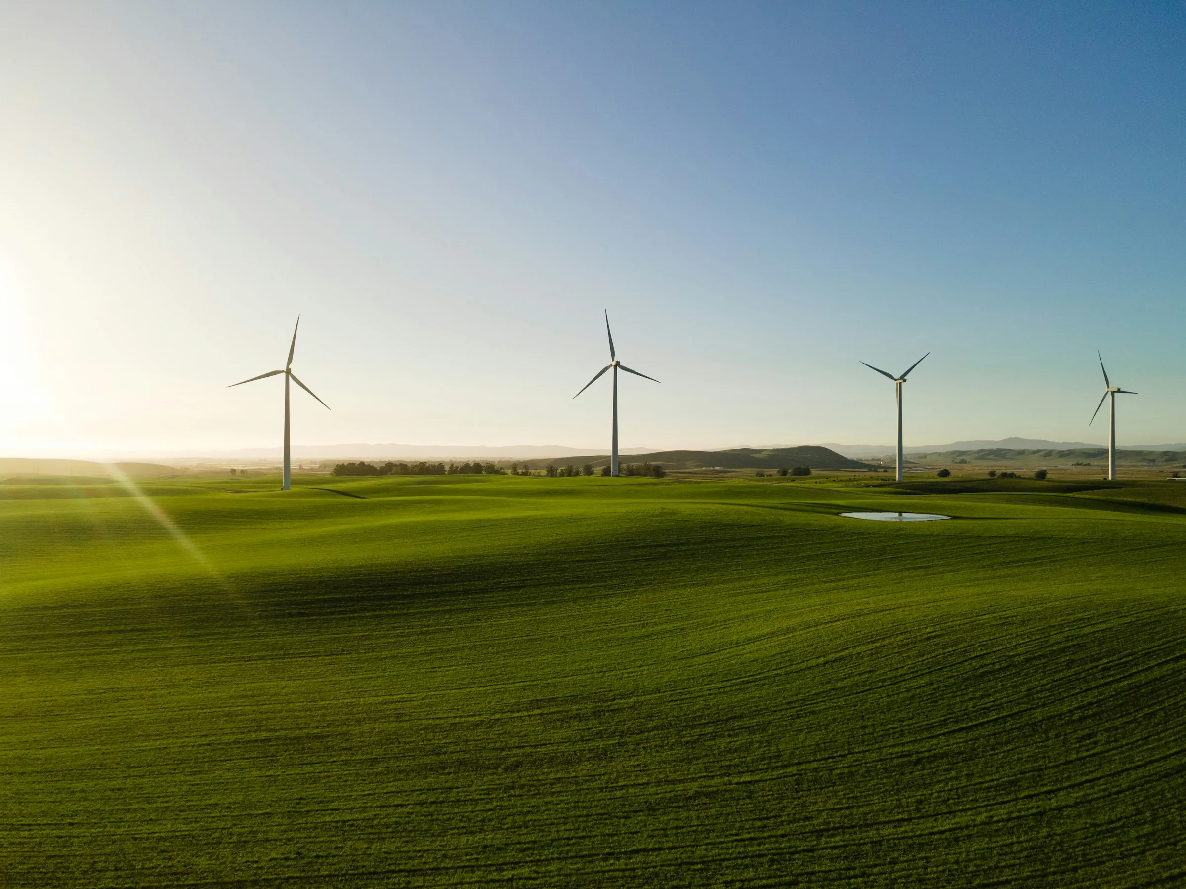 Four wind turbines standing on rolling green hills under a clear blue sky at sunrise or sunset.