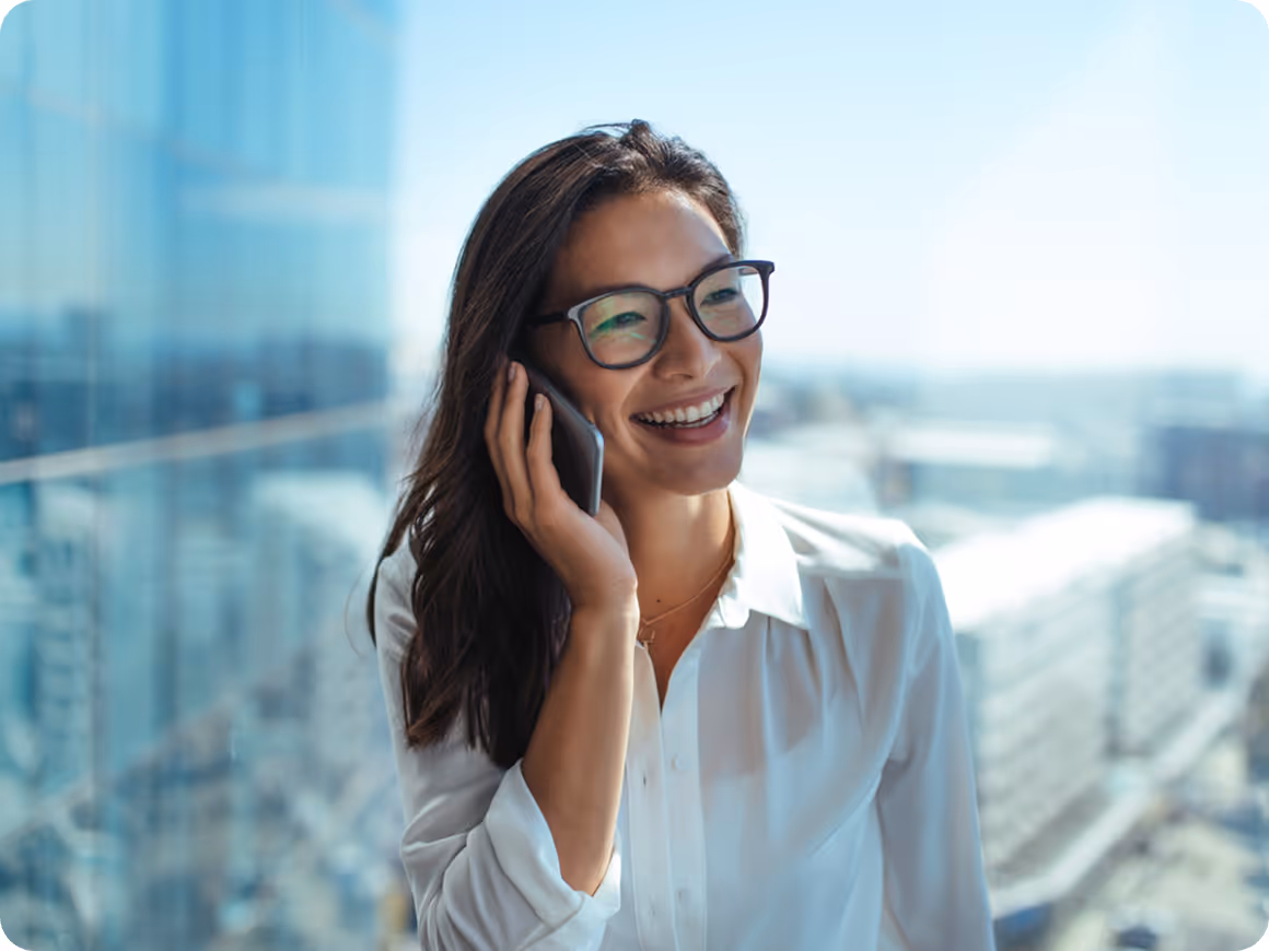 Jeune femme souriante portant des lunettes et une chemise blanche, parlant au téléphone avec un fond urbain flou.