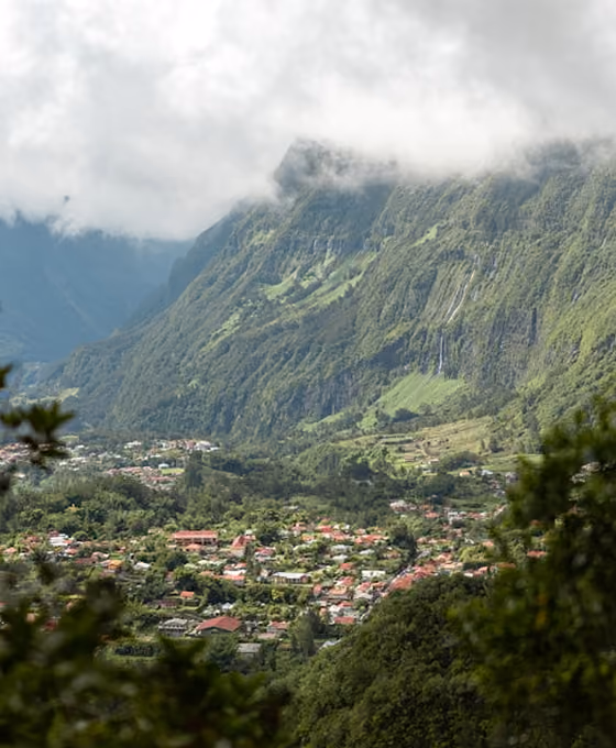 Village entouré de montagnes verdoyantes partiellement couvertes de nuages et de cascades fines.
