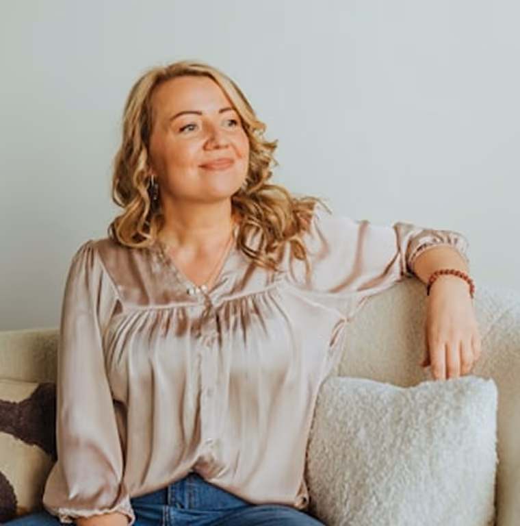 Femme souriante aux cheveux blonds bouclés assise sur un canapé beige avec un coussin blanc.