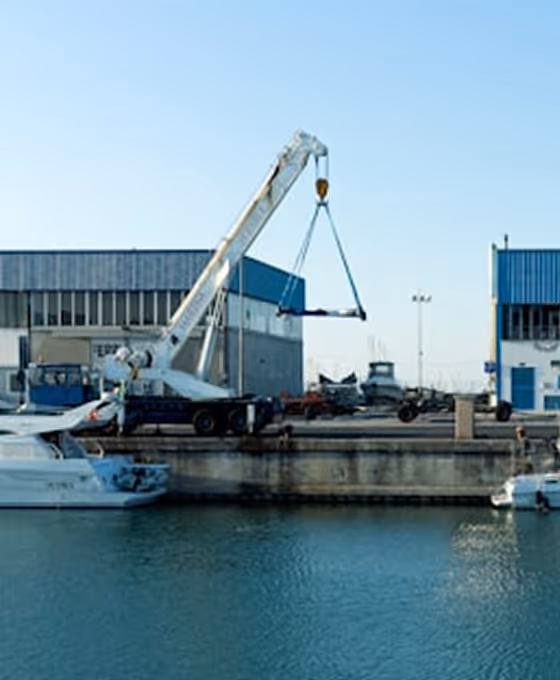 Grue mobile soulevant un bateau au port près quatre bâtiments industriels et deux bateaux amarrés.