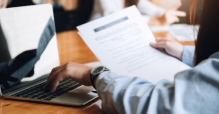 Personne tapant sur un ordinateur portable tout en tenant un document imprimé à une table.