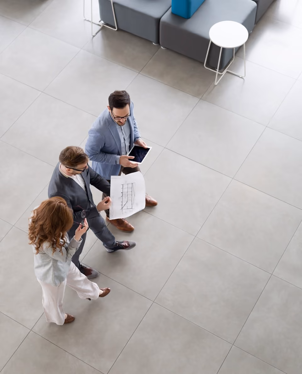Trois professionnels debout discutant autour d'un plan et d'une tablette dans un espace moderne.