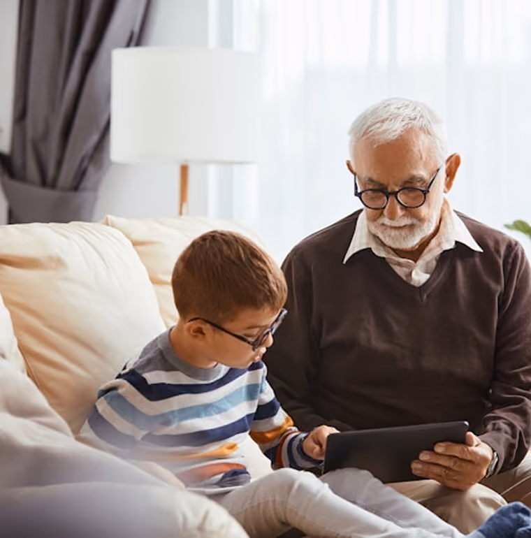 Un grand-père et son petit-fils regardent une tablette assis sur un canapé dans un salon lumineux.