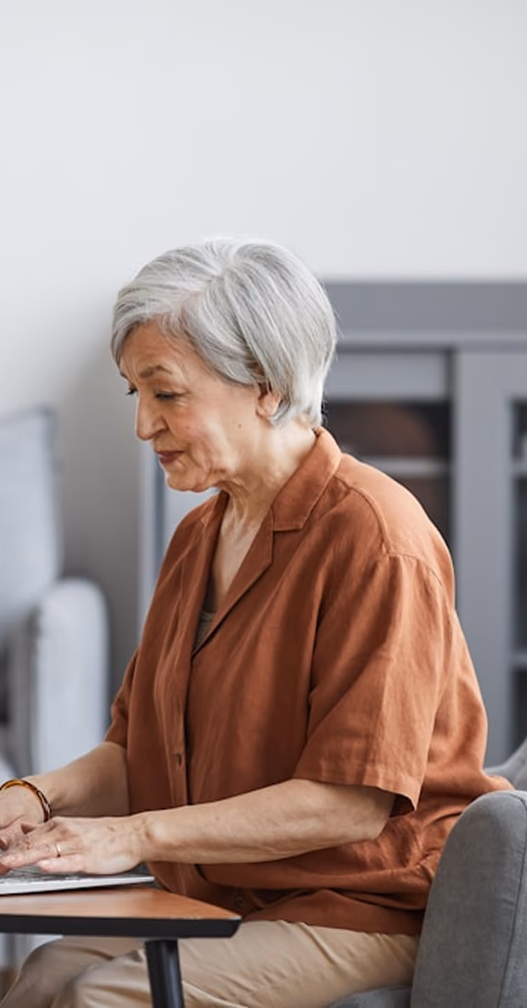 Femme âgée aux cheveux gris travaillant sur un ordinateur portable à une table intérieure.