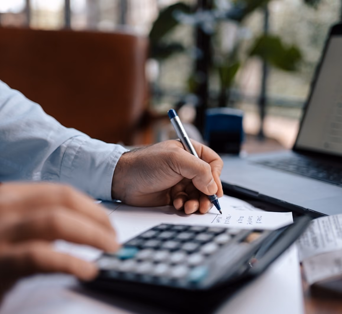 Une personne utilise une calculatrice et écrit des notes sur un document à côté d'un ordinateur portable.