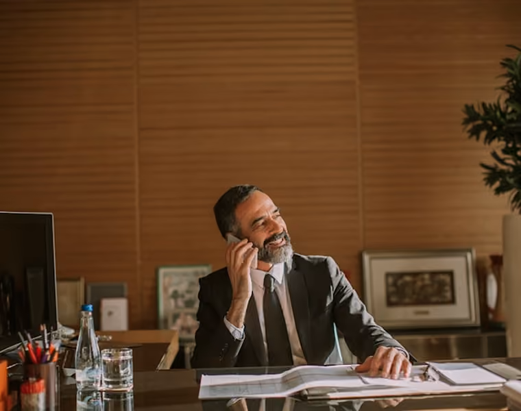Un homme d'affaires souriant parle au téléphone à son bureau avec des papiers et un verre d'eau.