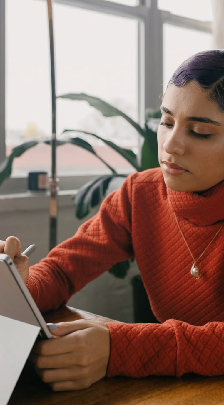 Femme concentrée en pull rouge écrivant sur une tablette avec un stylet près d'une fenêtre.