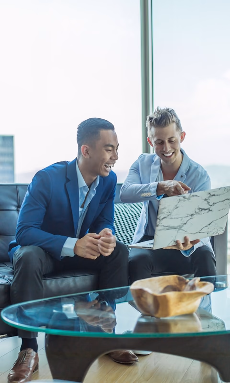 Deux hommes souriants regardent un ordinateur portable dans un bureau moderne avec vue sur la ville.