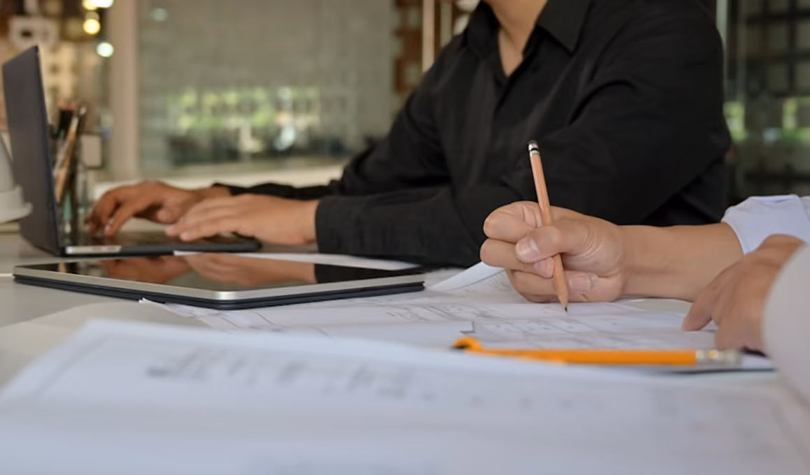 Deux personnes travaillant sur des plans avec crayon, tablette et ordinateur portable sur une table.