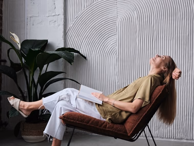 Femme détendue assise sur une chaise inclinée avec un livre ouvert sur ses genoux.