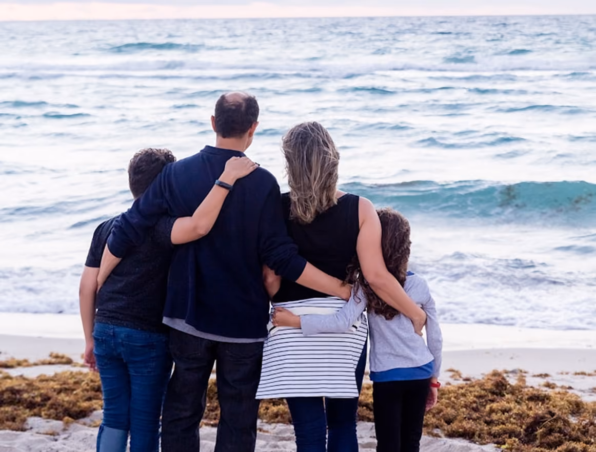 Famille de quatre personnes debout sur la plage, bras autour des épaules, regardant la mer.