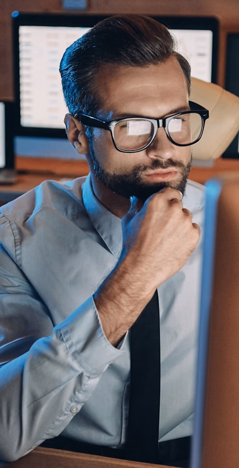 Homme d'affaires portant des lunettes, pensant devant un écran d'ordinateur avec des graphiques à l'arrière-plan.