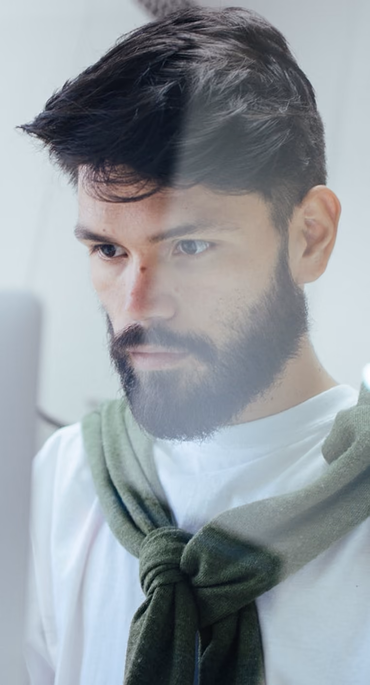 Homme barbu concentré regardant un écran, pull vert noué sur les épaules, fond clair flou.
