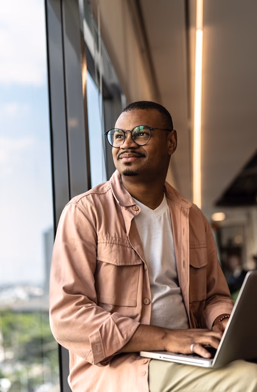 Homme portant des lunettes, souriant, utilisant un ordinateur portable près d'une grande fenêtre.