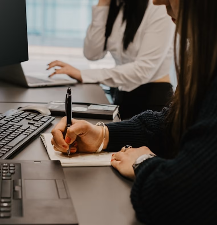 Deux femmes travaillent à un bureau avec ordinateurs, l'une prend des notes à la main.