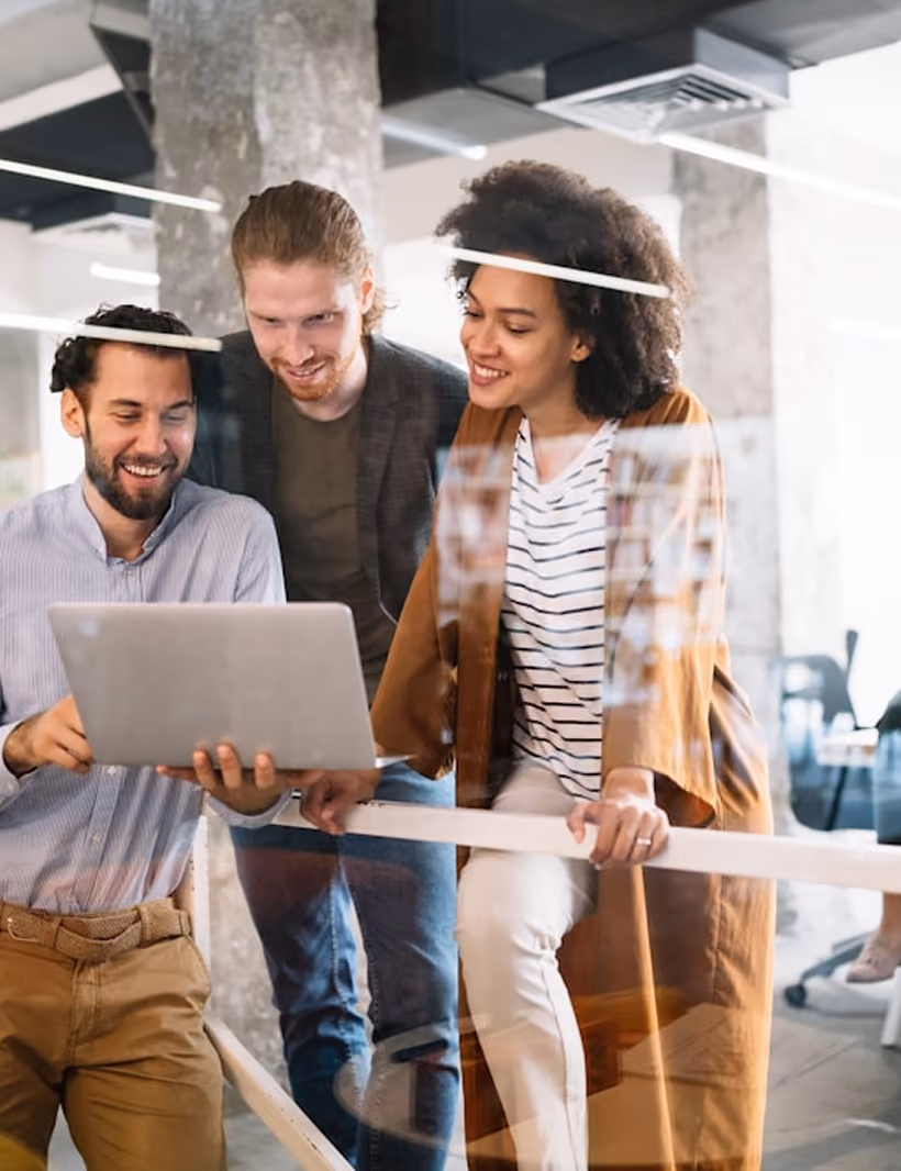 Trois collègues souriants regardent un ordinateur portable dans un bureau moderne.