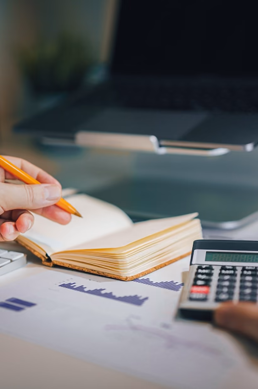 Person utilisant une calculatrice avec un carnet ouvert et des graphiques sur un bureau proche d’un ordinateur.