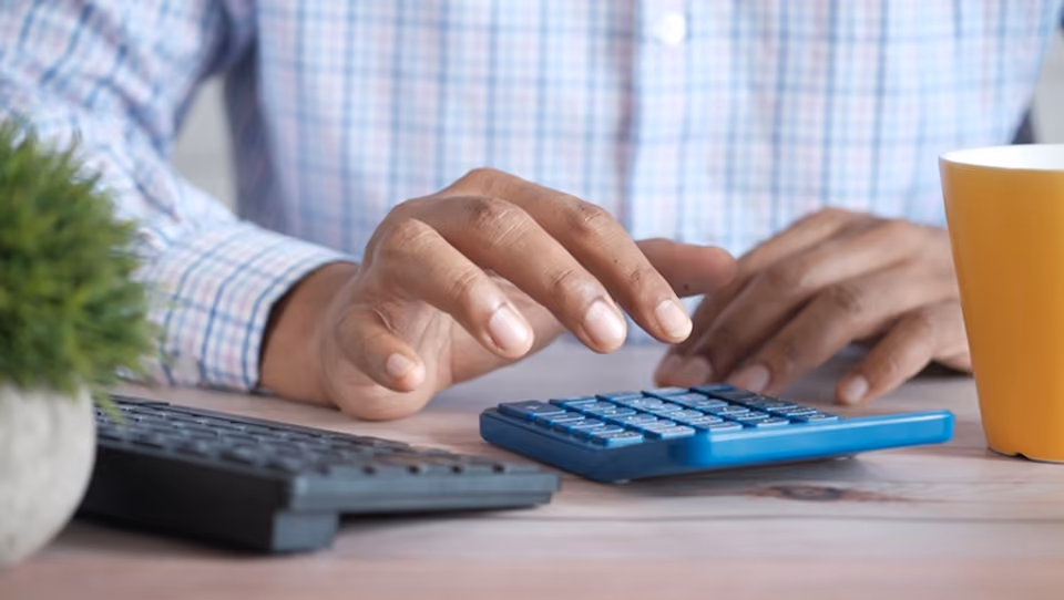 Une main utilise une calculatrice bleue sur un bureau avec un clavier et une tasse jaune à côté.