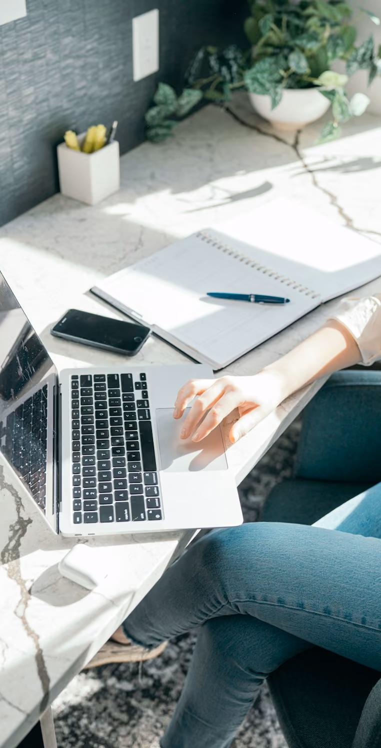 Person using a laptop on a marble desk with a phone, notebook, pen, and plant nearby.