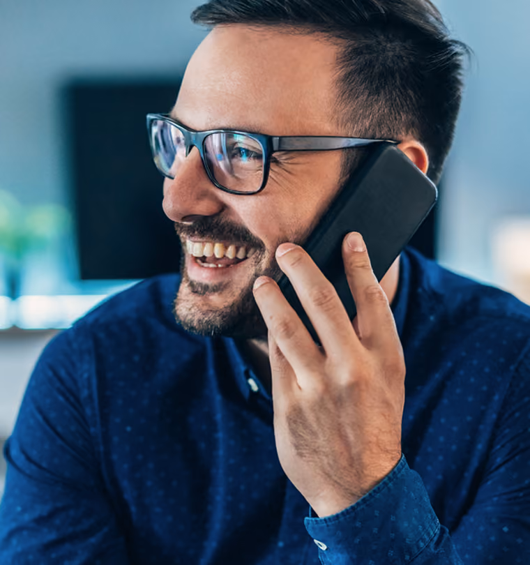 Homme souriant portant des lunettes parlant au téléphone avec un fond flou bleu.