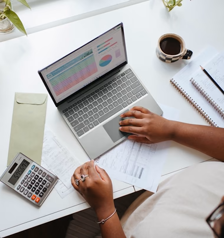 Personne travaillant sur un ordinateur portable avec calculatrice, papiers, carnet et tasse de café sur un bureau blanc.