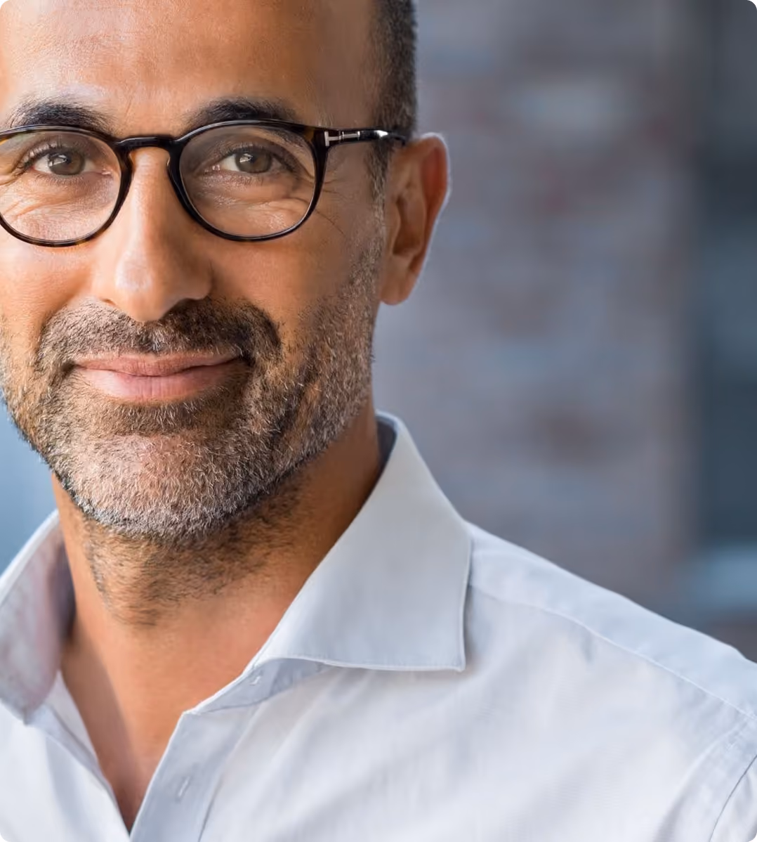 Homme souriant portant des lunettes noires et une chemise blanche, fond flou gris.