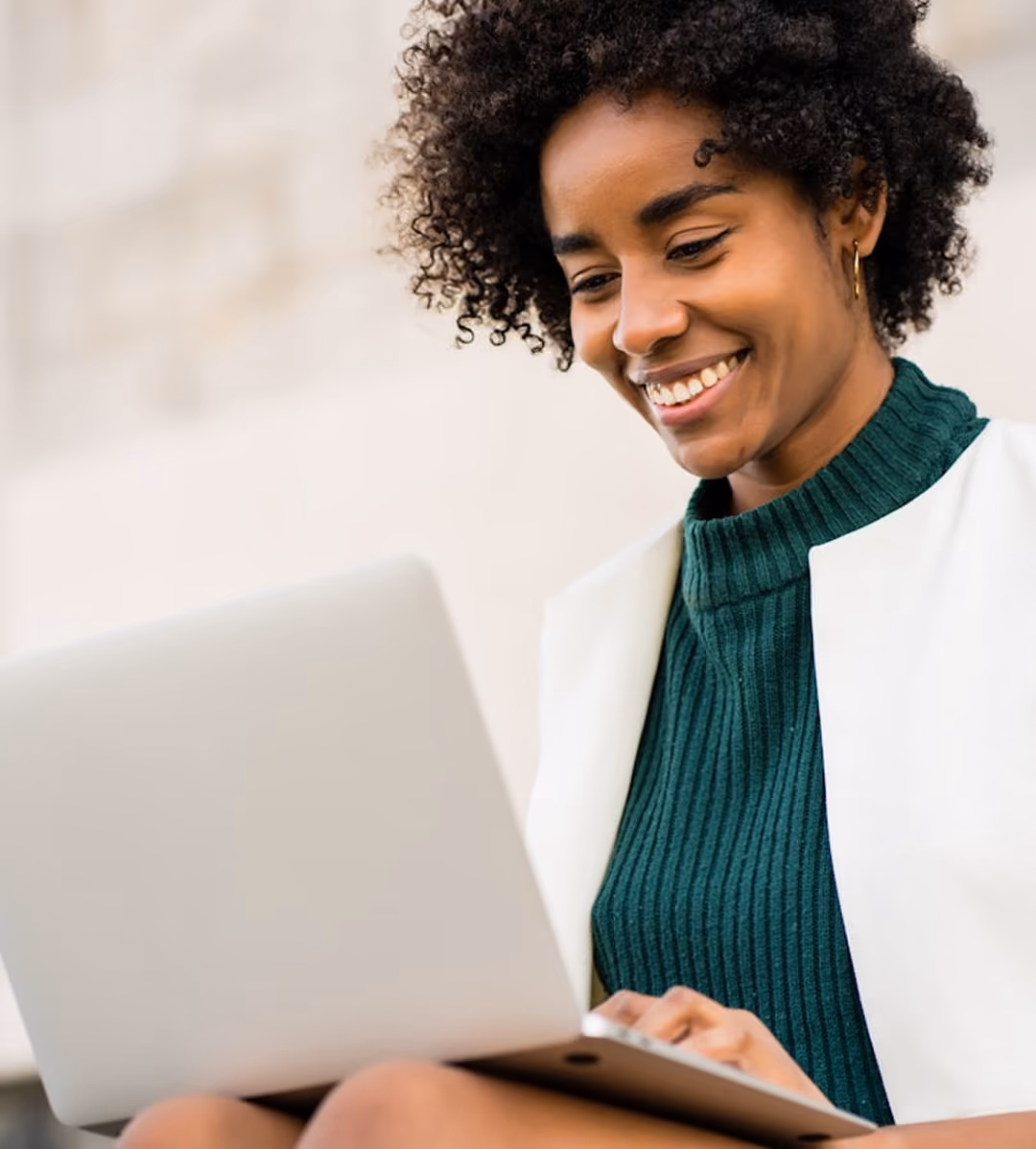 Jeune femme souriante utilisant un ordinateur portable, vêtue d'un pull vert et d'une veste blanche.