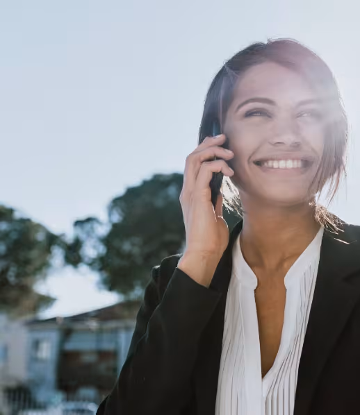 Femme souriante parlant au téléphone en extérieur avec un ciel clair et des arbres en arrière-plan.