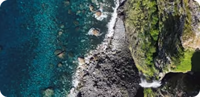 Vue aérienne d'une côte avec eau turquoise, rochers sombres et végétation verte dense.