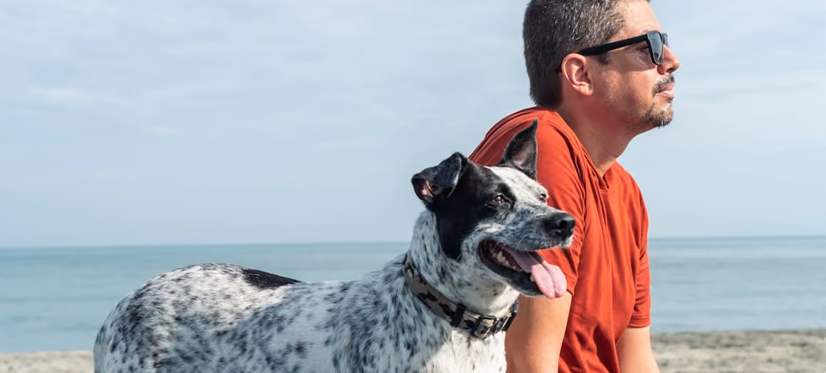 Homme en t-shirt rouge et lunettes de soleil assis à côté d'un chien blanc tacheté à la plage.