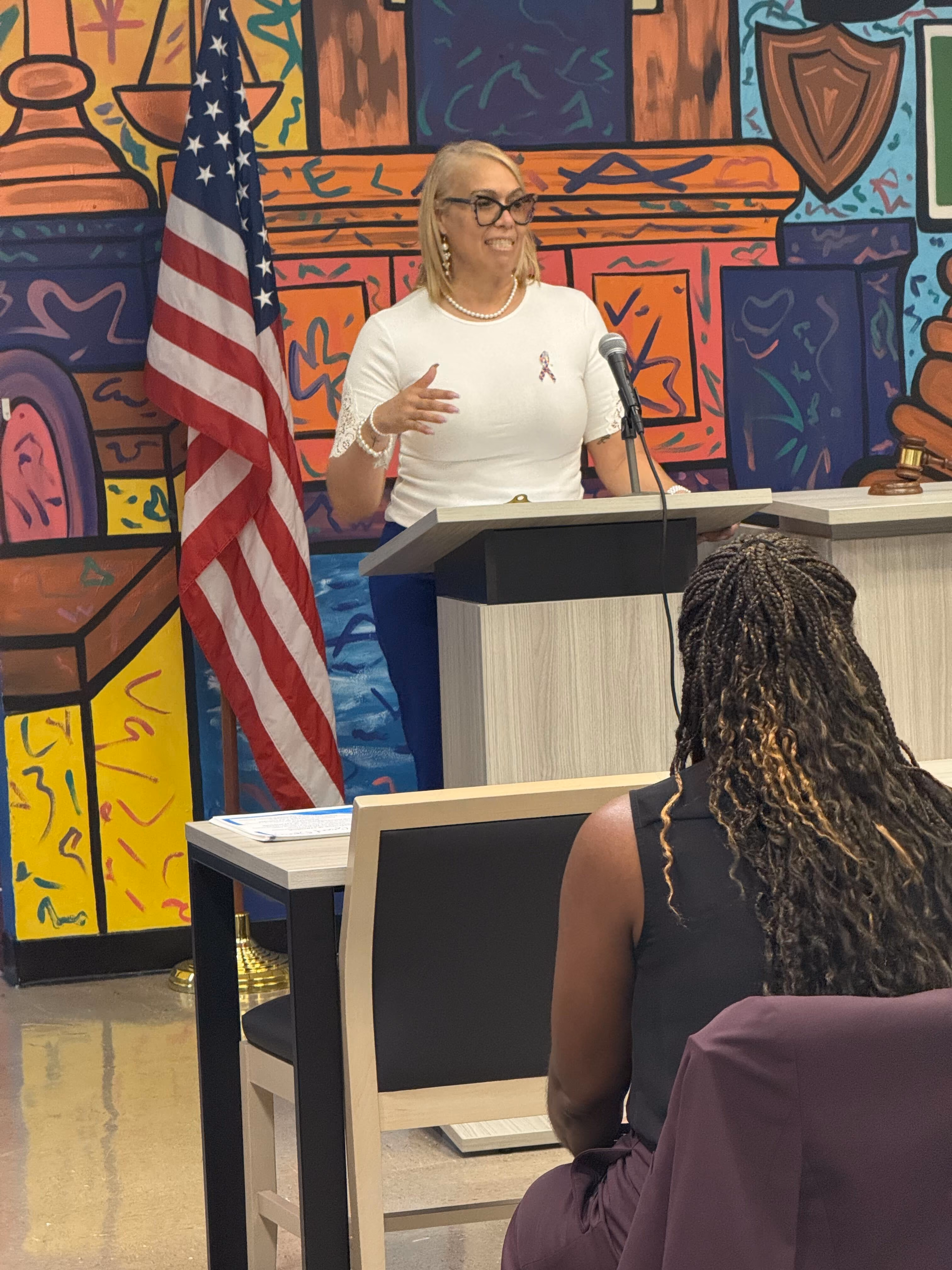 Woman speaking at podium with American flag and colorful mural