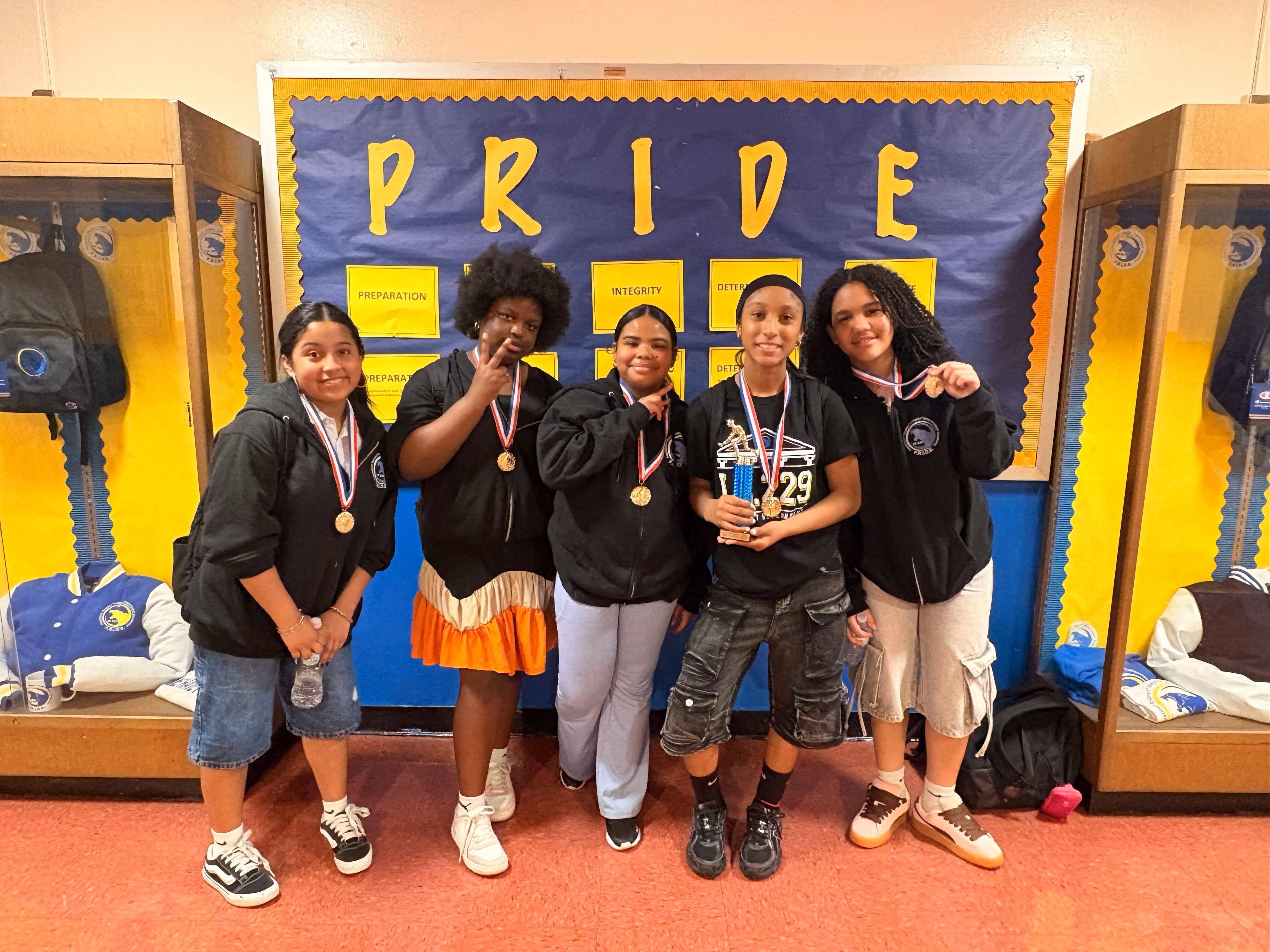 Five students displaying medals and trophy in front of Pride bulletin board