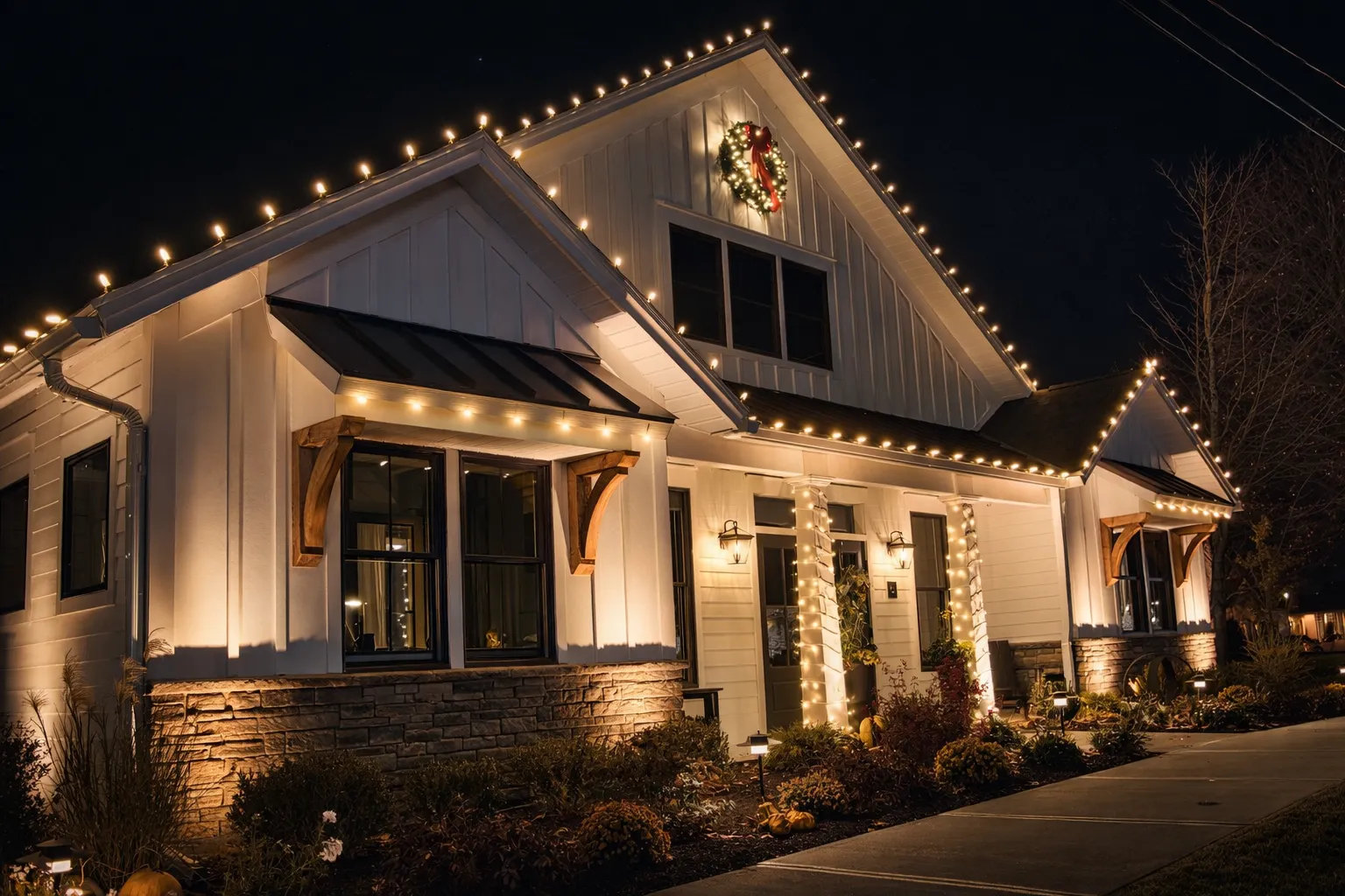 White house decorated with warm white string lights along roof edges and wrapped around porch columns at night, featuring a lit Christmas wreath above a window.