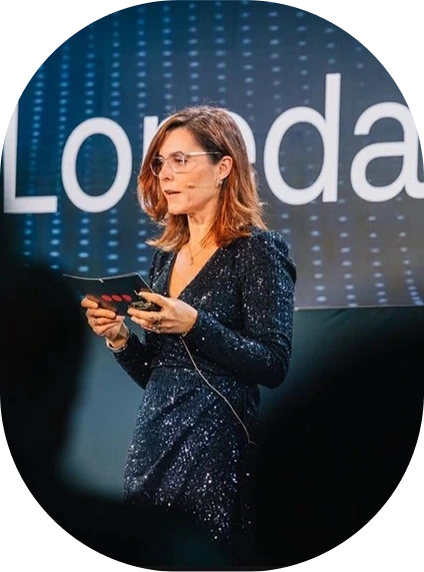 Woman in a dark blue sequined dress speaking on stage with a microphone headset and holding note cards.
