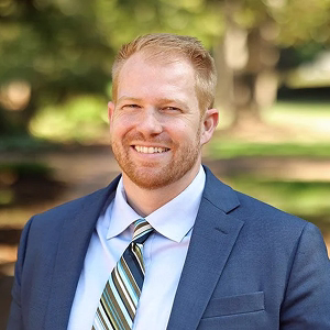 Smiling man with short light brown hair and beard wearing a blue suit, striped tie, and light blue shirt outdoors.