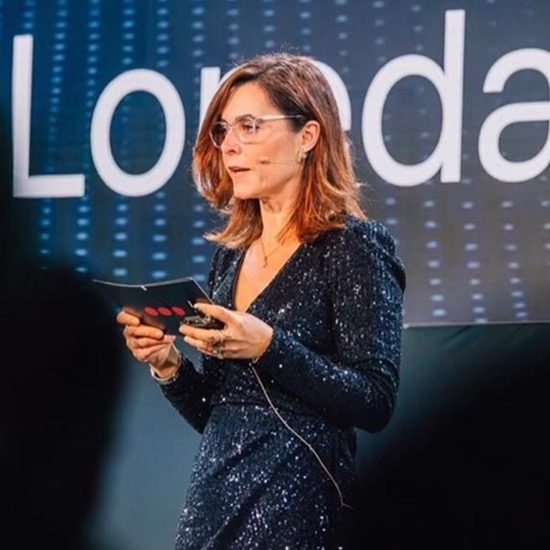 Woman with glasses speaking on stage holding a card, wearing a sparkly dark dress in front of a screen displaying text.