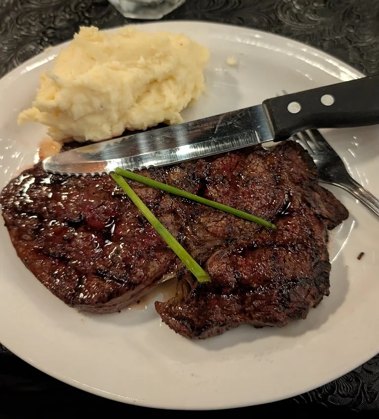 Grilled steak with two green chives on top alongside mashed potatoes on a white plate with a knife and fork.