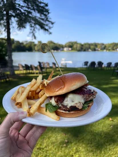 Hand holding a plate with a cheeseburger topped with lettuce and grilled onions, served with French fries, with a lake and chairs in the background.