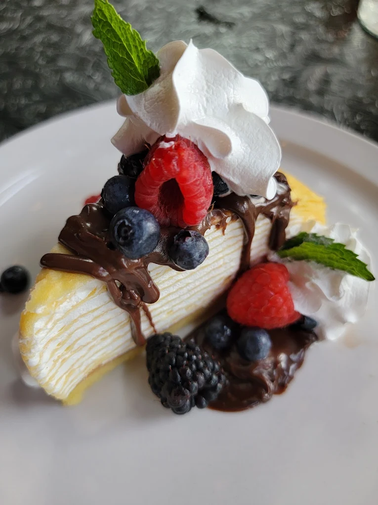 Slice of layered crepe cake topped with blueberries, raspberries, chocolate drizzle, whipped cream, and mint leaves on a white plate.