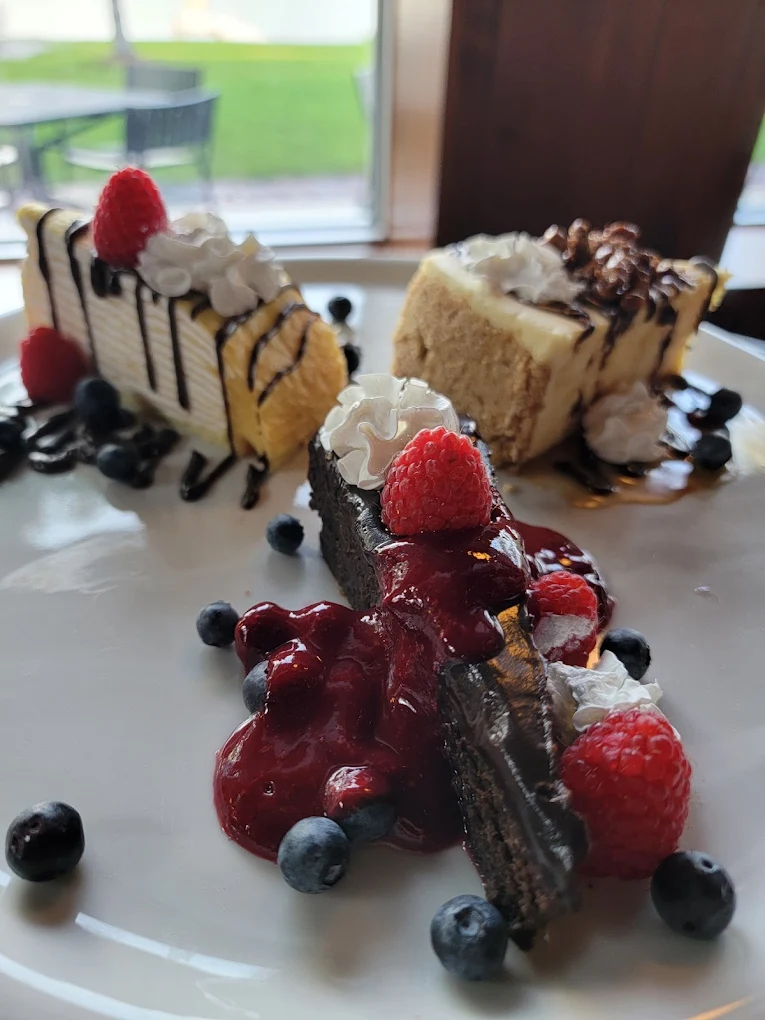 Three assorted dessert slices on a white plate, garnished with whipped cream, raspberries, blueberries, chocolate drizzle, and berry sauce.