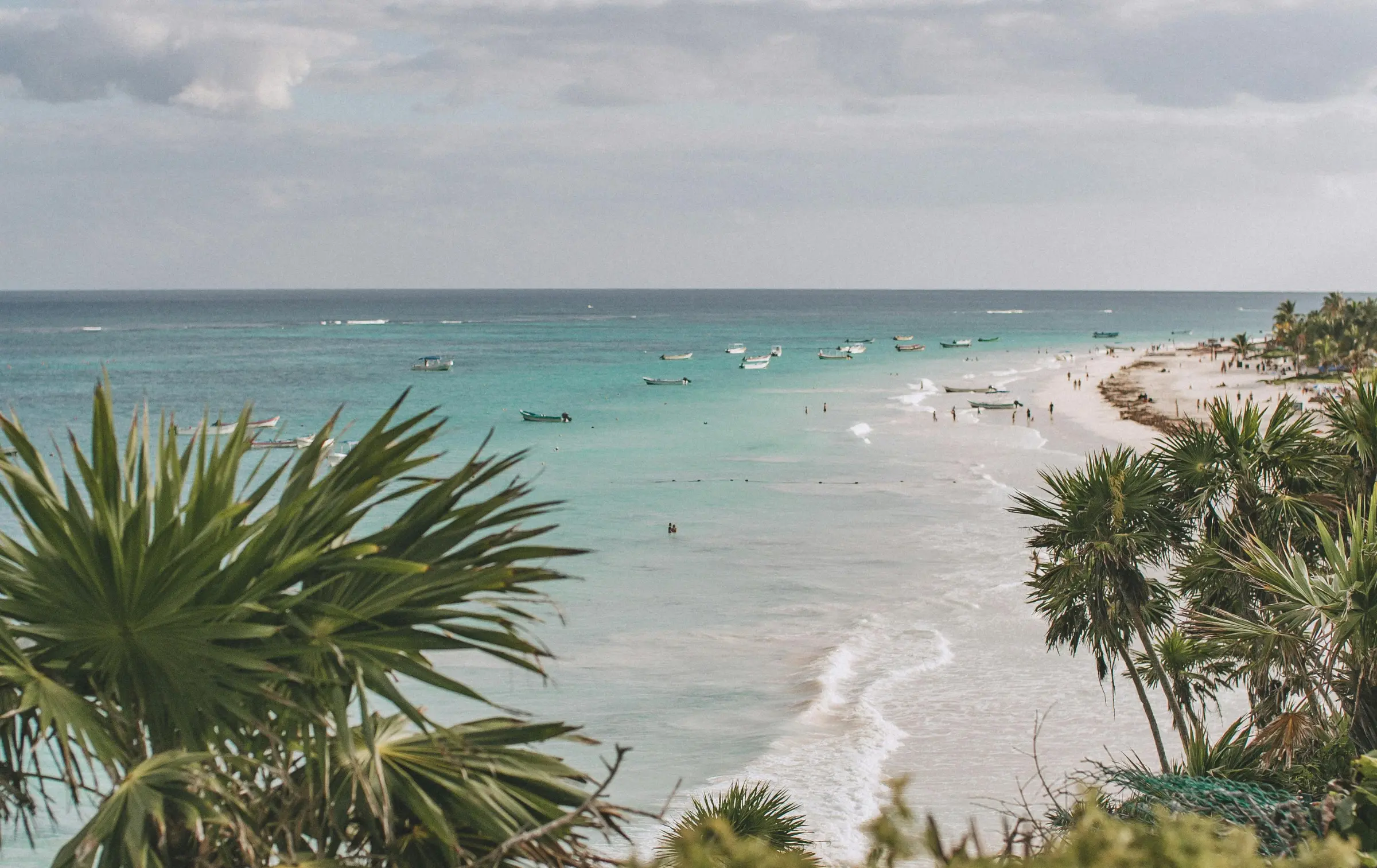 Turquoise ocean with small boats near a sandy beach lined with palm trees under a cloudy sky in Tulum, Mexico.