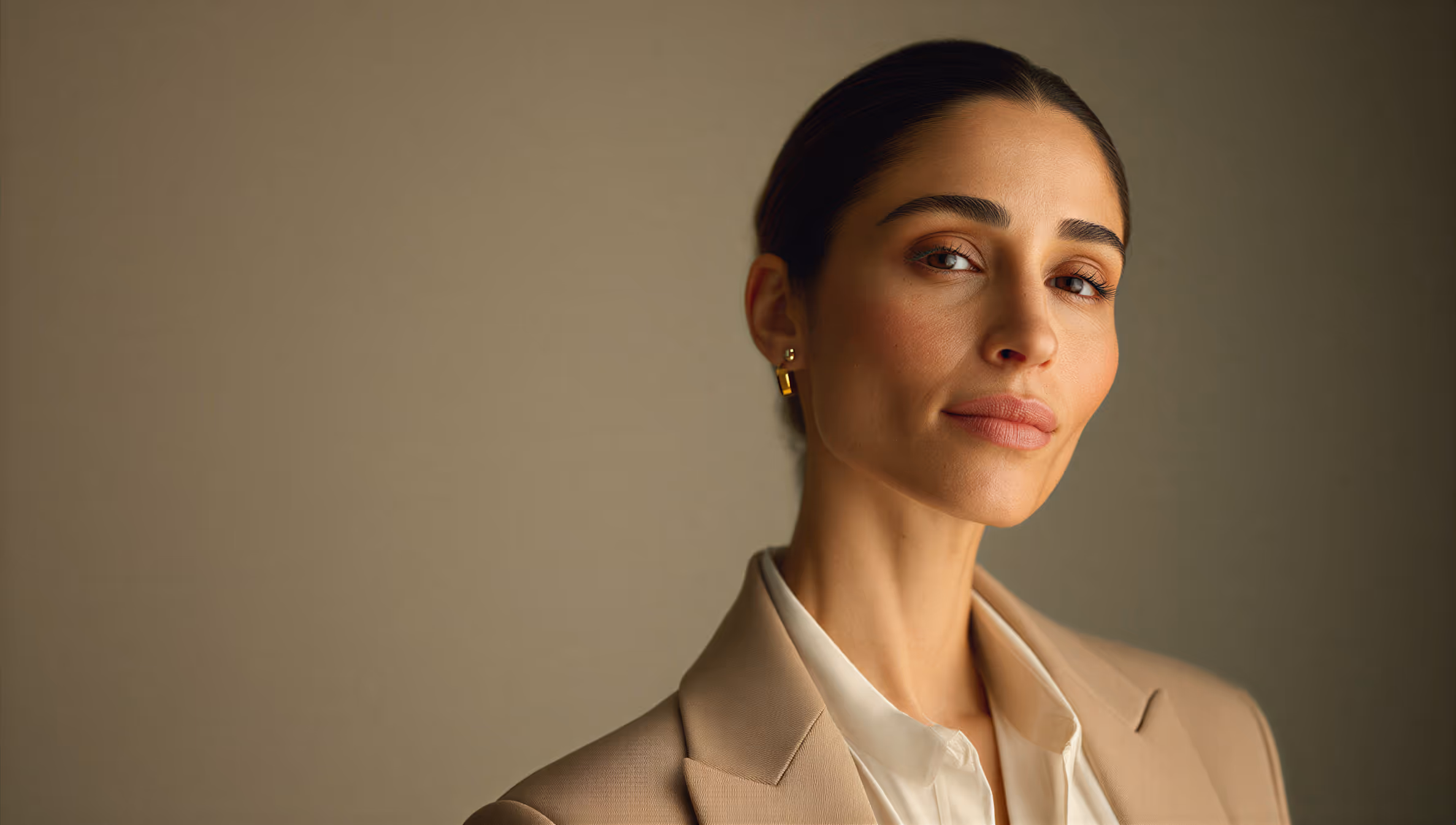 Confident woman with dark hair pulled back wearing a beige blazer and white blouse against a neutral background.