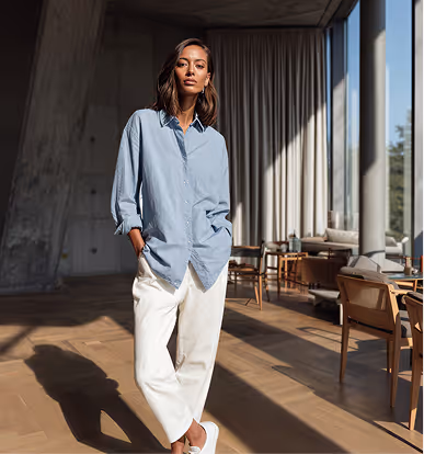 Woman wearing a light blue button-up shirt and white pants standing in a sunlit modern room with large windows and wooden furniture.