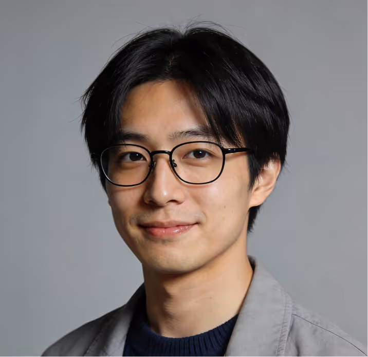Portrait of a young man with black hair and glasses smiling slightly against a gray background.