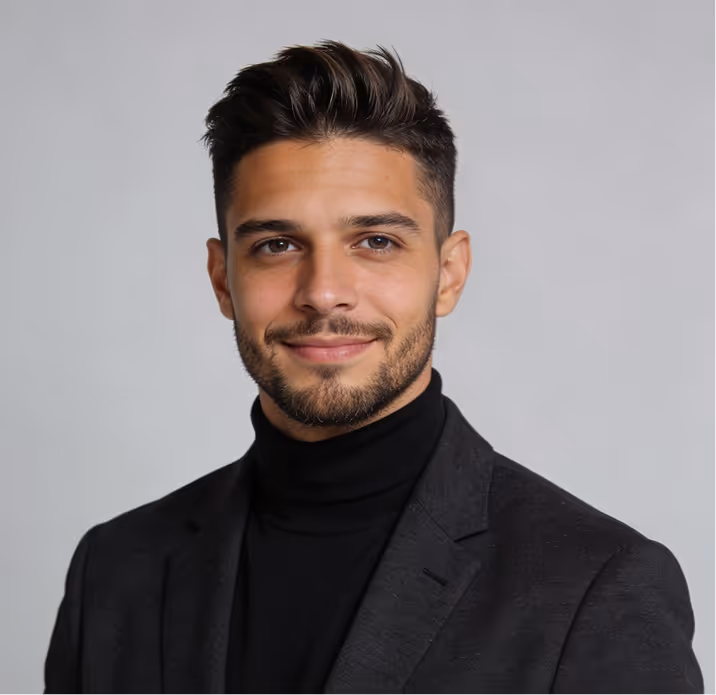 Smiling young man with short dark hair and beard wearing a black turtleneck and dark blazer against a gray background.