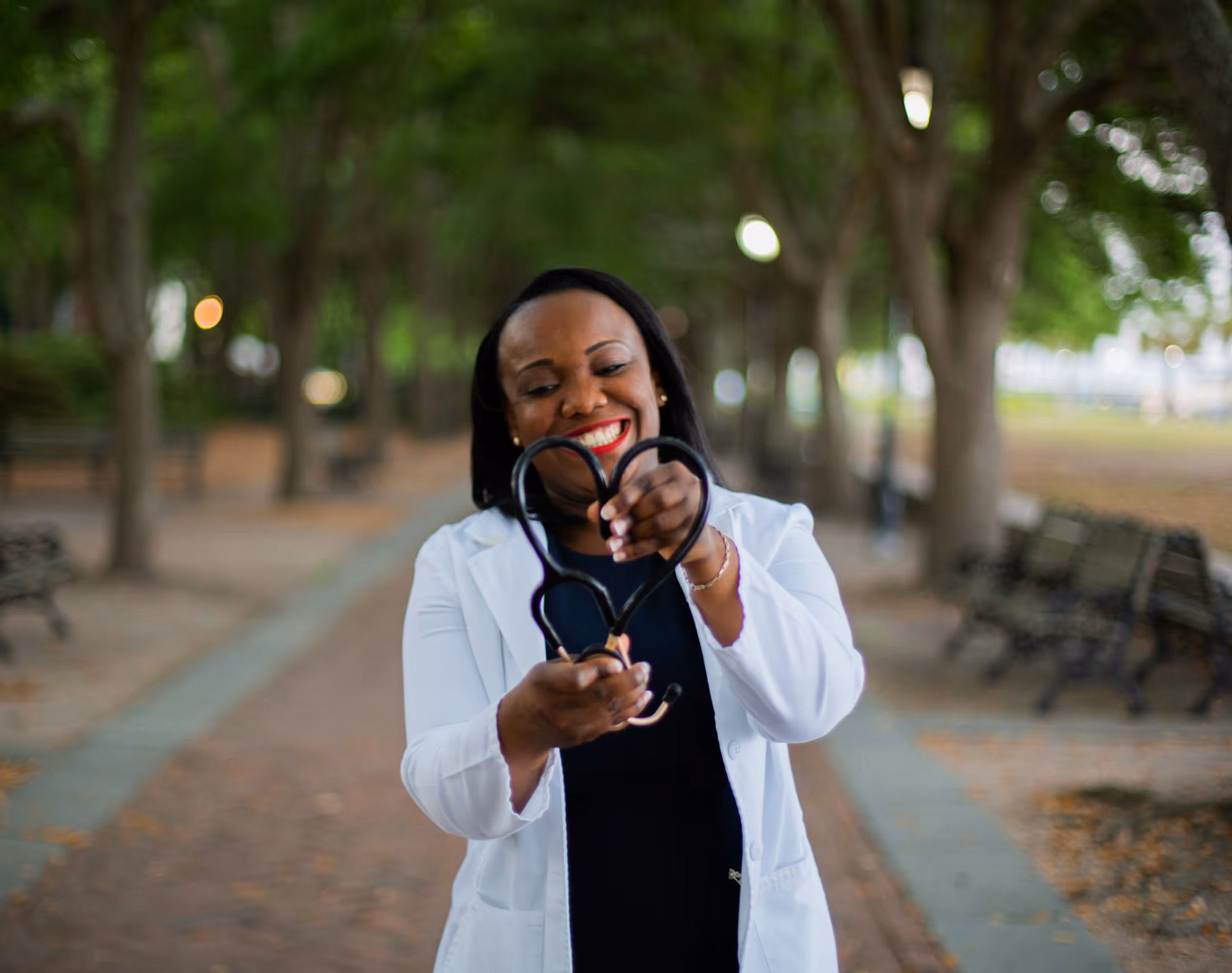 A Black female healthcare provider, wearing a white lab coat over a black top, stands smiling and holding the tubing of her stethoscope bent into the shape of a heart. She is standing outdoors on a brick pathway lined with trees, symbolizing the dedication and heart independent providers bring to their communities.