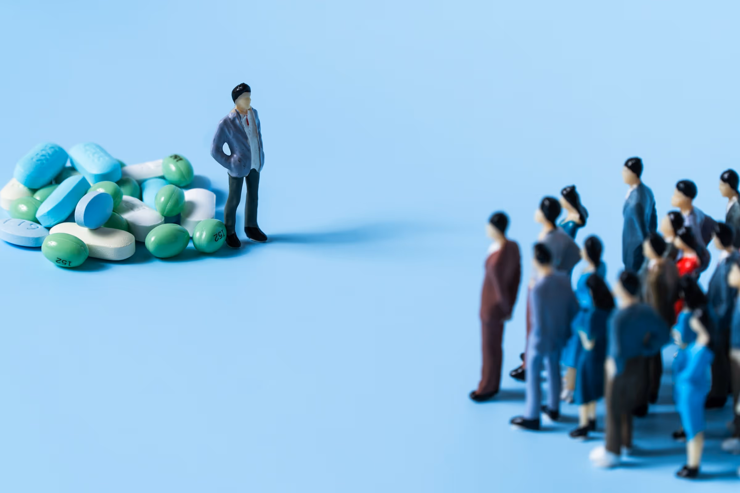 A small plastic figurine of a person in a suit stands beside a large pile of blue, green, and white pills, facing a larger crowd of miniature figurines. The image uses the pills and the crowd to symbolize the US healthcare system and its independent providers.