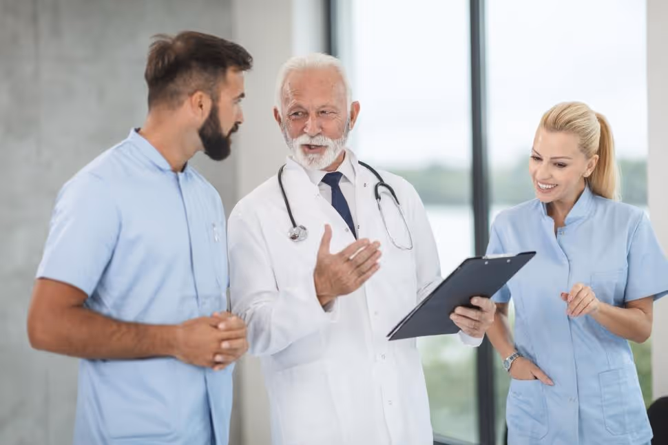 An older, experienced male doctor in a white coat is speaking and gesturing to a younger male nurse and a female nurse or administrator, all wearing scrubs or professional attire. They are reviewing a document on a clipboard. The image symbolizes the critical discussions and planning among staff needed to address potential Medicaid cuts.